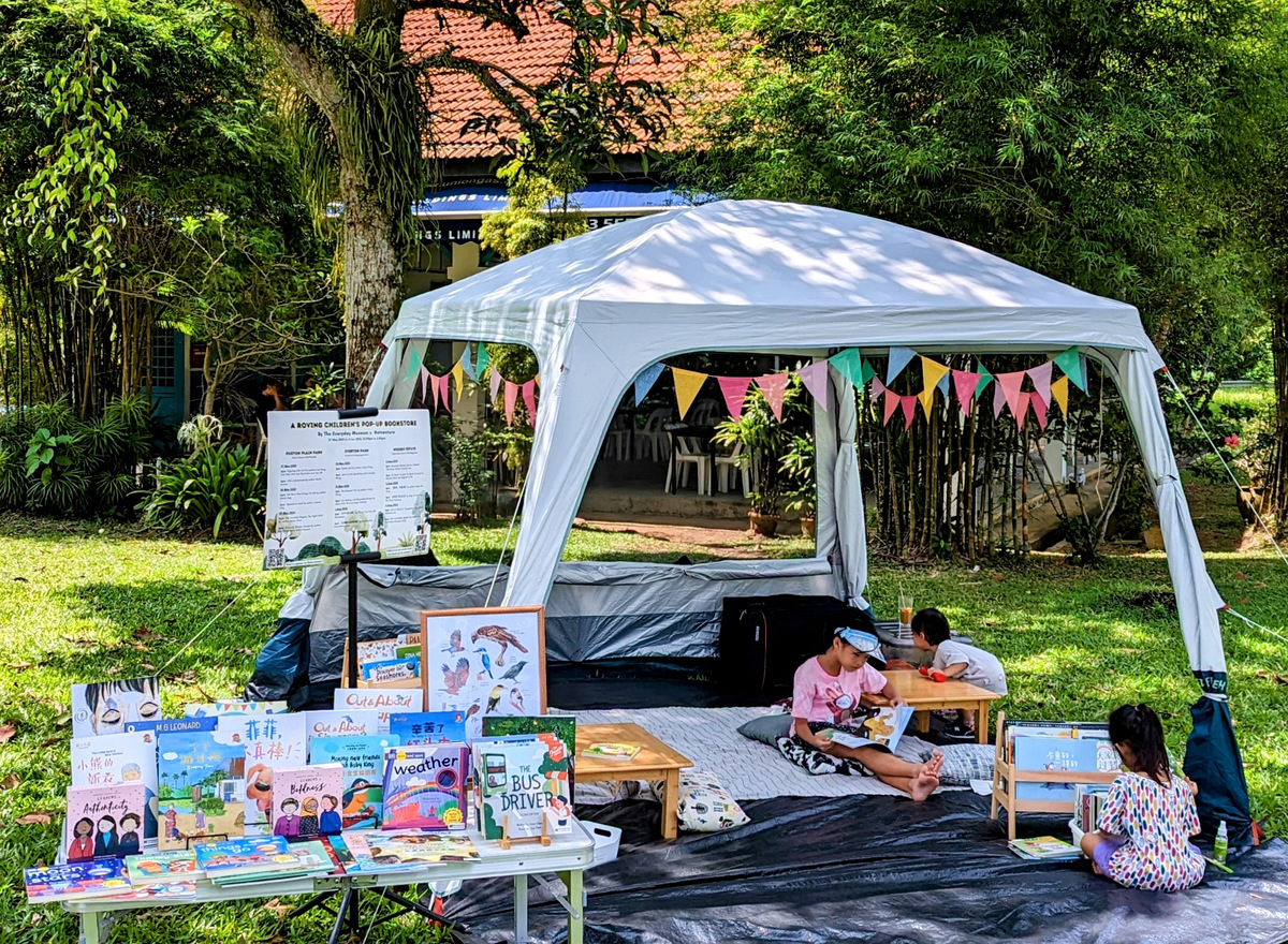 Natventure pop-up booth: a table of Singapore children's books in front of a backdrop of Cecilia reading to children